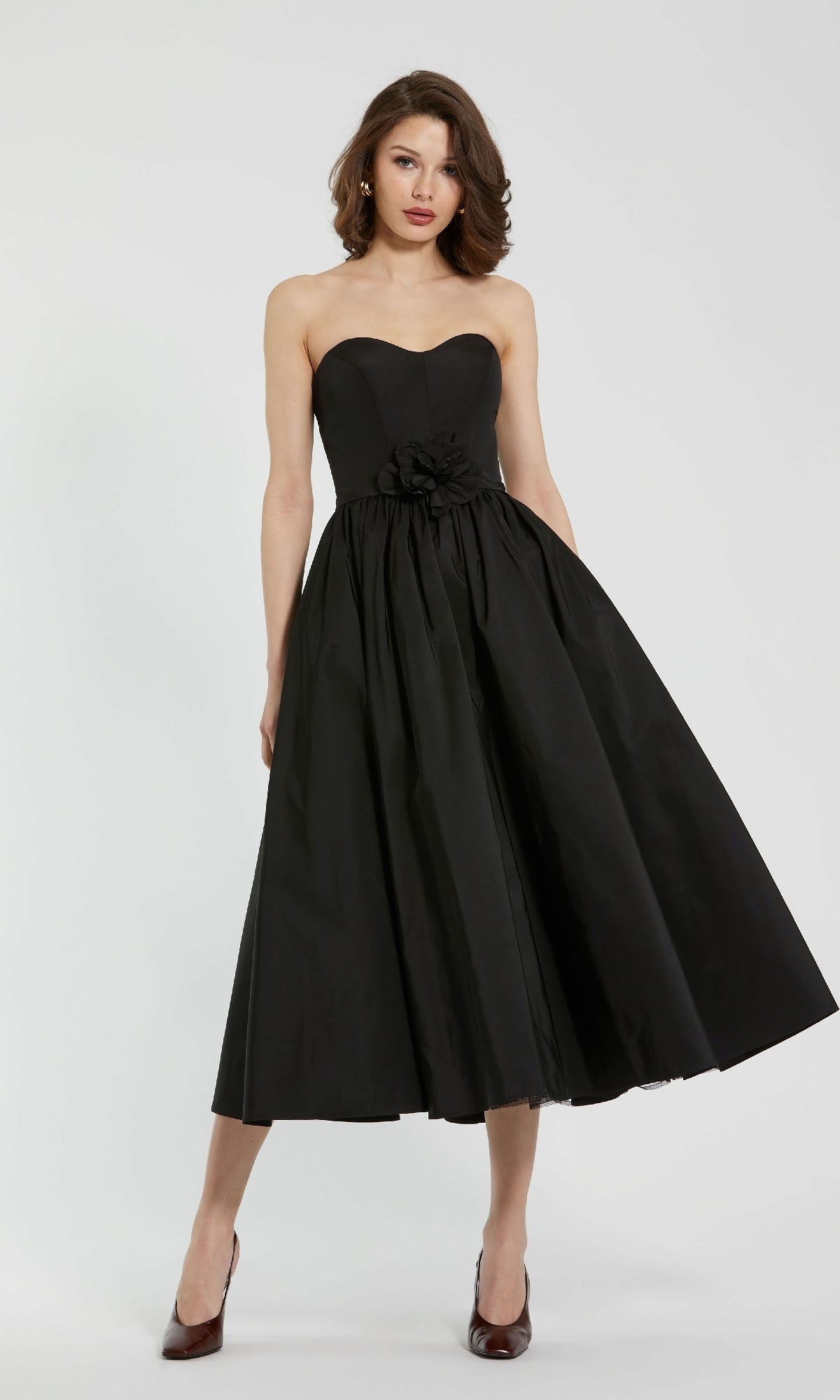 A woman stands against a plain background wearing the Mac Duggal 21023 Midi Strapless Semi-Formal Dress, featuring a fitted bodice and a floral accent at the waist.